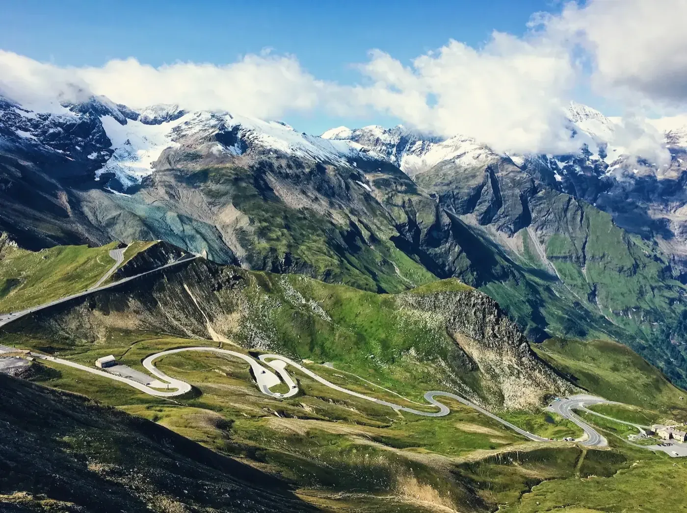 Hairpin road winding through a green alpine valley beneath snow-capped peaks and a glacier.