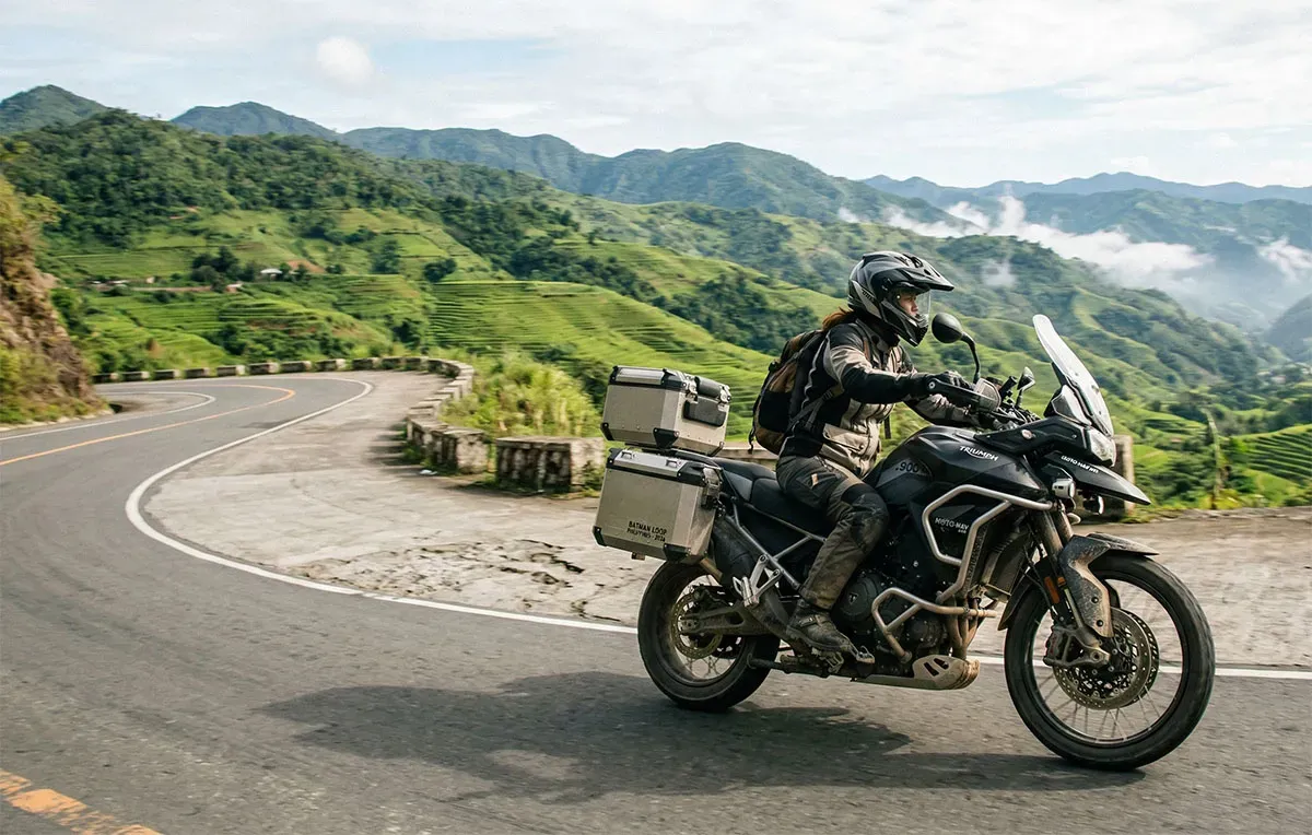 A rider on a loaded adventure motorcycle rounding a bend above green rice terraces.