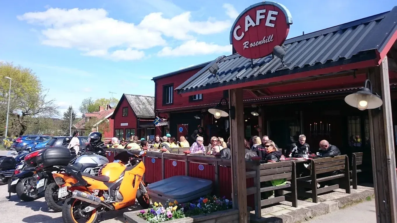 Motorcyklar parkerade framför Café Rosenhills röda byggnad och fullsatta uteservering en solig dag.