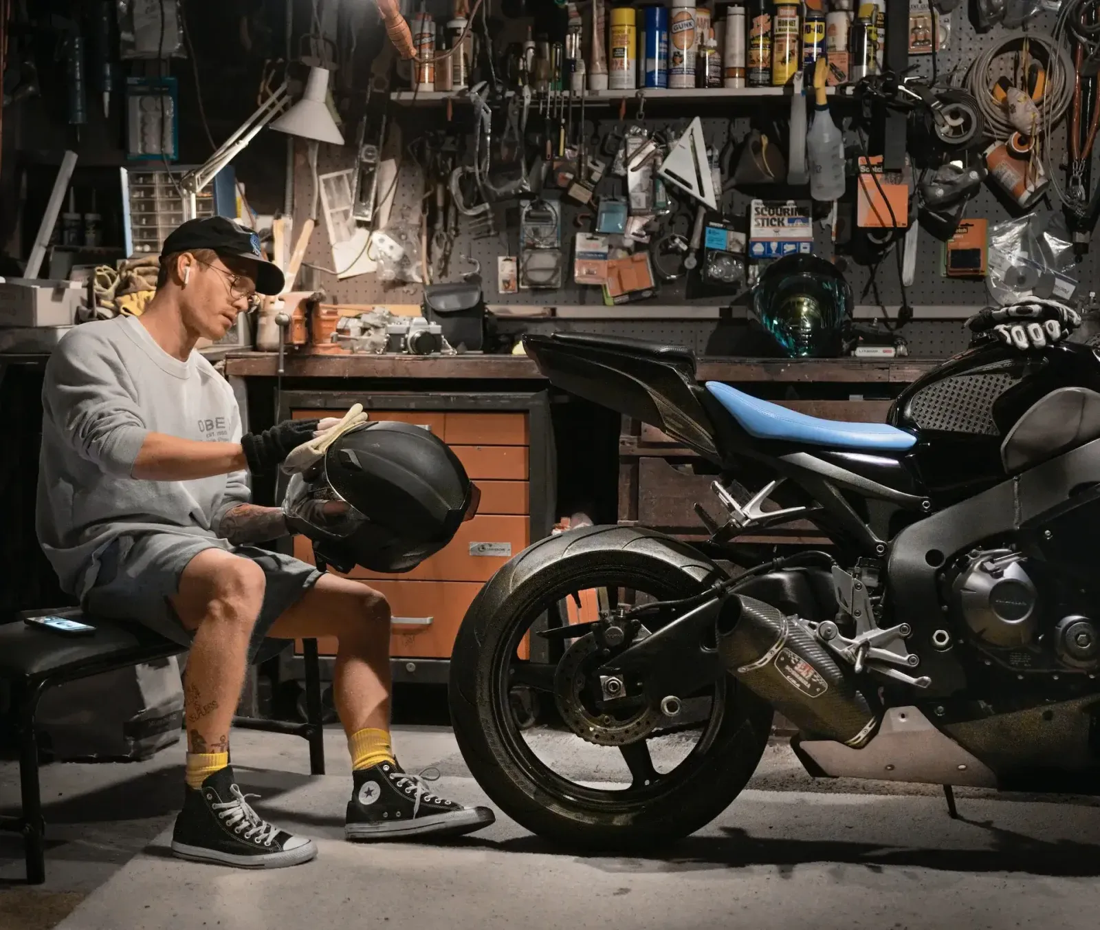 A rider wiping a helmet in a garage workshop beside a black sport motorcycle.