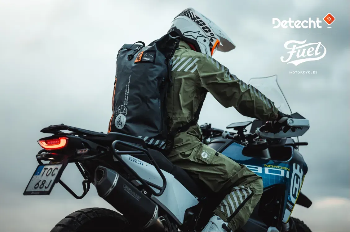 A rider in green gear with a backpack on an adventure motorcycle, beside Detecht and Fuel Motorcycles logos.
