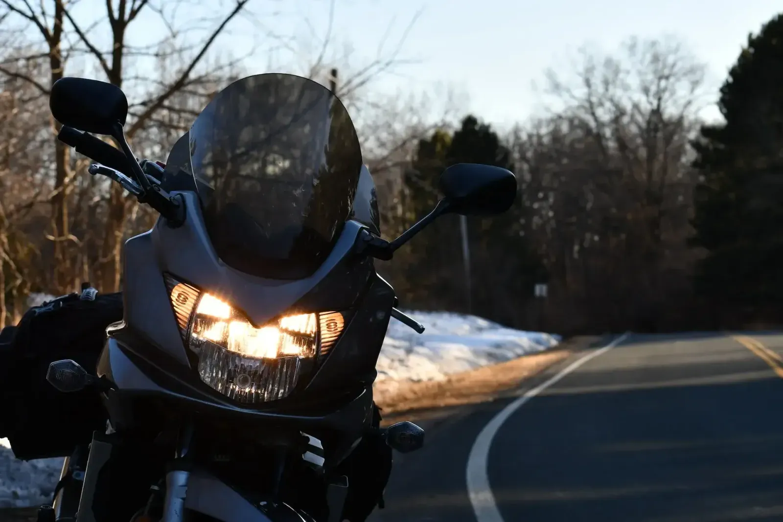 Motorcycle with its headlight on, parked beside a quiet road with patches of snow along the verge.