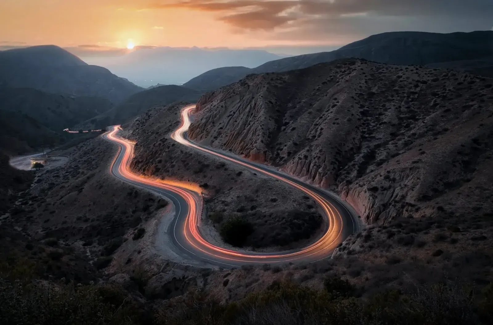 Light trails from passing vehicles tracing a winding mountain road at sunset.