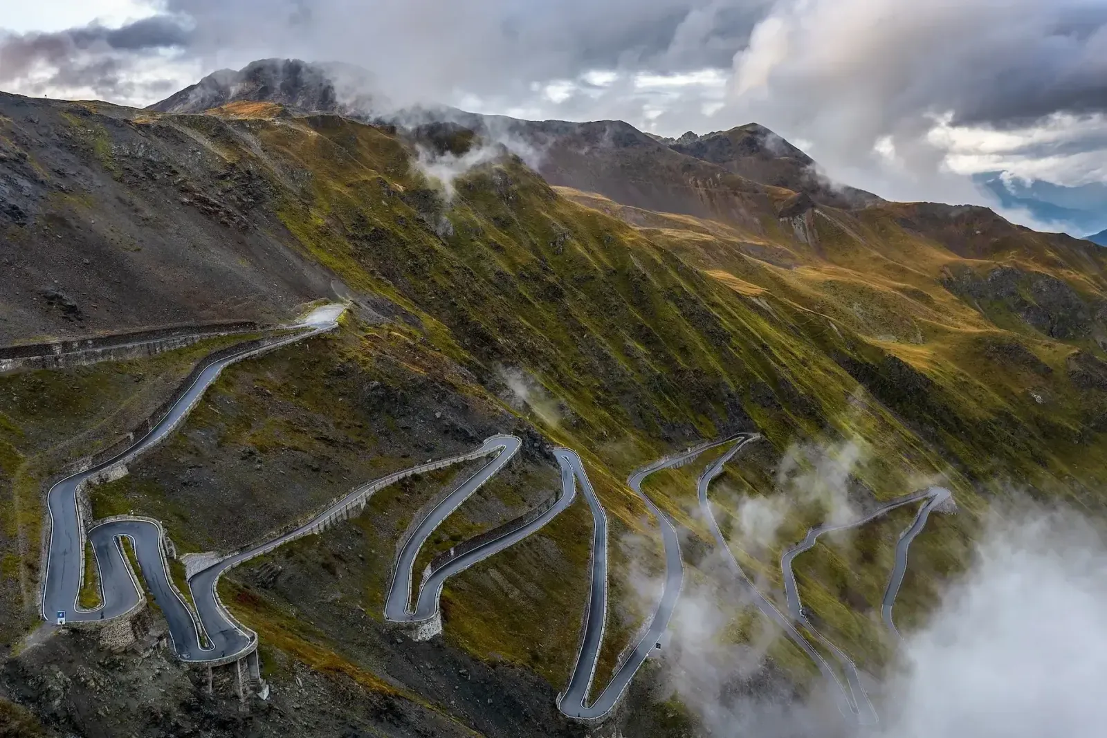 Hairpin bends of the Stelvio Pass zigzagging up a green mountainside beneath low clouds.