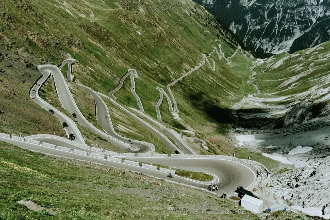 Motorcycles and cars climbing the stacked hairpin bends of the Stelvio Pass up a steep valley.