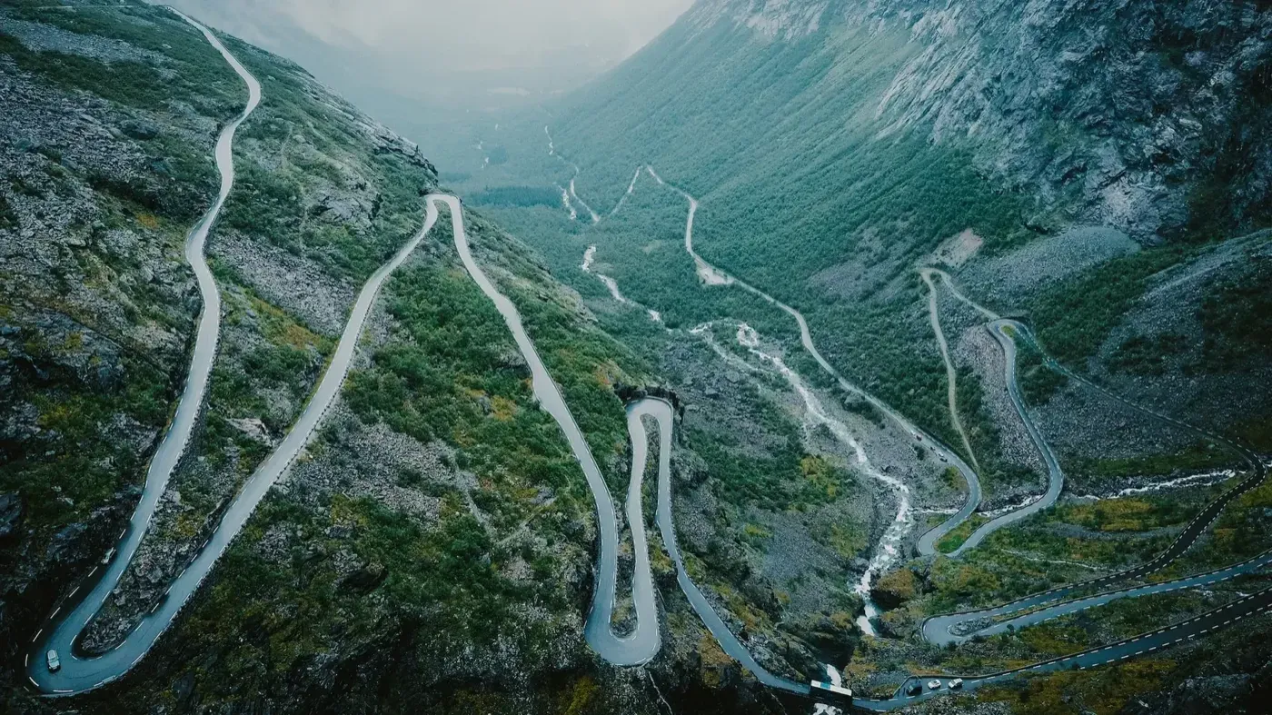 Aerial view of the Trollstigen road zigzagging down a steep green mountainside beside a waterfall.